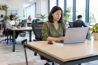 Professional woman with a walking stick at her desk in a bright modern office, working on a laptop with co-workers in the background.